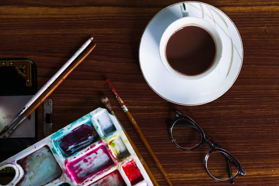 Overhead view of ceramic mug of hot drink on wooden table near paint palette and eyewear